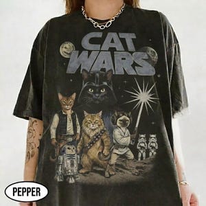 Może przedstawiać: Ciemnoszara koszulka z grafiką "Cat Wars". Projekt przedstawia koty przebrane za postacie z Gwiezdnych Wojen, z kotem Darth Vader w centrum. Koszulka ma wygląd vintage.