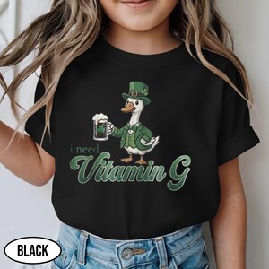 May include: Black t-shirt featuring a cartoon goose in a green suit and hat, holding a beer mug with a shamrock. The text "i need Vitamin G" is displayed in green. The word "BLACK" is in a white oval at the bottom.