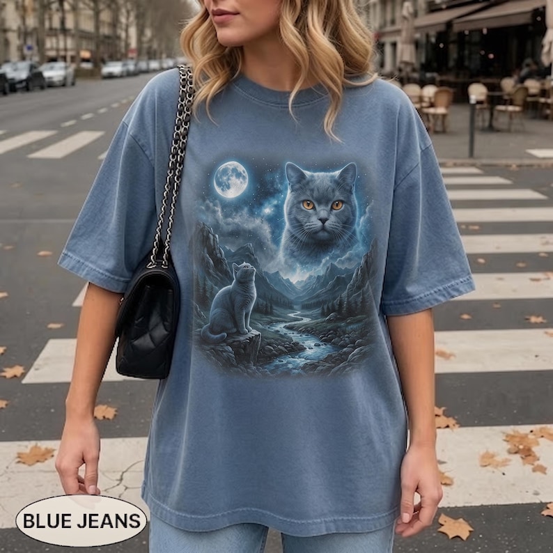 Pode incluir: Uma t-shirt de manga curta em azul poeirento com uma estampa gr&aacute;fica do rosto de um gato grande no c&eacute;u noturno acima de um gato menor sentado em uma rocha perto de um riacho de montanha e uma lua cheia.