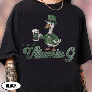 May include: Black t-shirt featuring a cartoon goose in a green suit and hat, holding a beer mug with a shamrock. The text "i need Vitamin G" is written in green script below the goose. The word "BLACK" is in a white oval at the bottom.