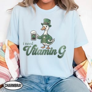 May include: Light blue t-shirt featuring a cartoon duck wearing a green suit and hat, holding a beer mug with a shamrock. The text "i need Vitamin G" is below the duck. The shirt has a crew neck.
