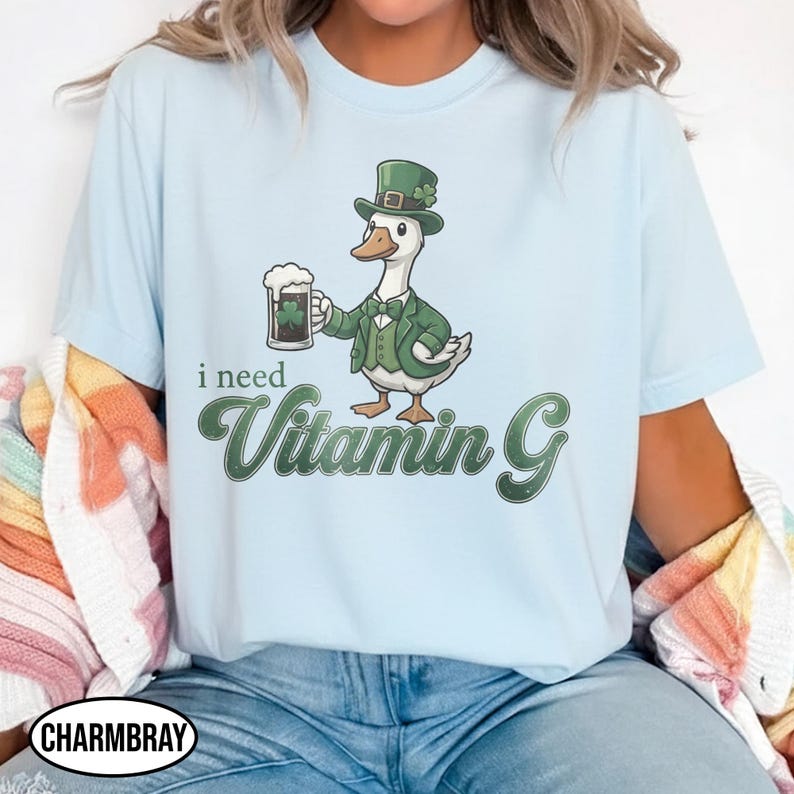 May include: Light blue t-shirt featuring a cartoon duck wearing a green suit and hat, holding a beer mug with a shamrock. The text "i need Vitamin G" is below the duck. The shirt has a crew neck.