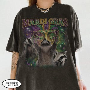 May include: Dark gray t-shirt with a Mardi Gras theme. The shirt features a graphic of three raccoons, a mask, and the words "MARDI GRAS" in purple and gold. The raccoons are set against a background of purple, green, and gold confetti.