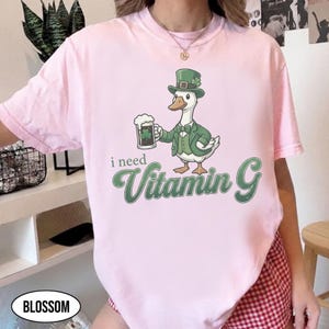 May include: Light pink t-shirt featuring a cartoon goose in a green suit and hat, holding a beer mug with a shamrock. The text "i need Vitamin G" is printed in green. The shirt is a casual wear item.