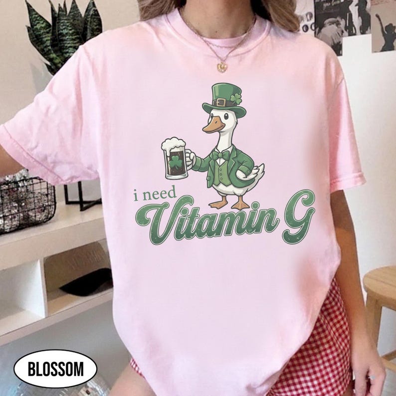 May include: Light pink t-shirt featuring a cartoon goose in a green suit and hat, holding a beer mug with a shamrock. The text "i need Vitamin G" is printed in green. The shirt is a casual wear item.