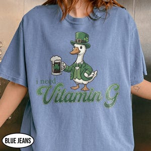 May include: Blue t-shirt featuring a cartoon duck in a green suit and hat, holding a beer with a shamrock. The text "i need Vitamin G" is printed below the duck in green script.