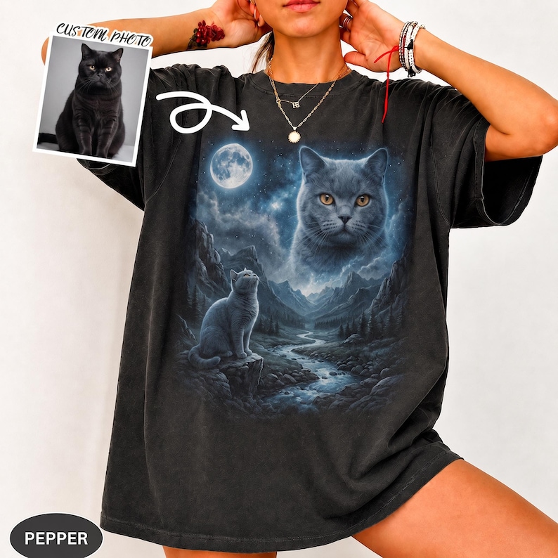 Pode incluir: Uma t-shirt preta com uma estampa gr&aacute;fica de um gato sentado num penhasco rochoso sobre um rio numa paisagem montanhosa sob uma lua cheia, com o rosto de um gato grande no c&eacute;u. Uma pequena foto mostra um gato preto com o texto CUSTOM PHOTO.