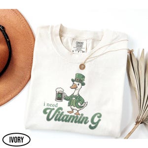 May include: Ivory-colored t-shirt featuring a cartoon goose in a green suit and hat, holding a beer with a shamrock. The text "i need Vitamin G" is printed in green. A gold necklace and a brown hat are also visible.