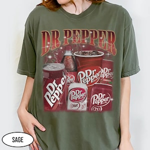 May include: Sage green t-shirt featuring a Dr Pepper graphic. The design includes the Dr Pepper logo, cans, a bottle, and a cup of the soda. The text "Dr Pepper" is displayed prominently at the top of the graphic.