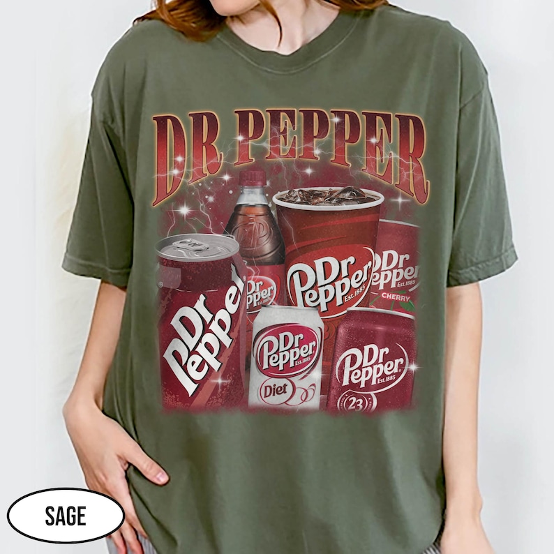 May include: Sage green t-shirt featuring a Dr Pepper graphic. The design includes the Dr Pepper logo, cans, a bottle, and a cup of the soda. The text "Dr Pepper" is displayed prominently at the top of the graphic.