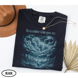 Puede incluir: Camiseta negra con un gráfico de un dinosaurio en un cielo tormentoso, un gallo y el texto "Remember who you are". También se ven un collar dorado y un sombrero marrón.