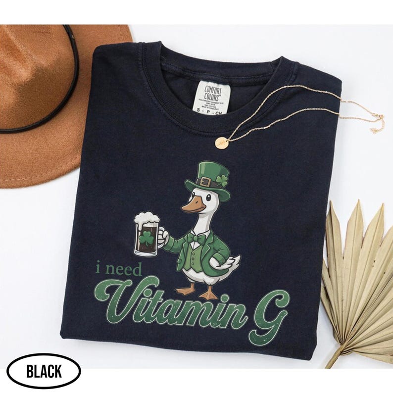 May include: Black crew neck t-shirt with a graphic of a goose wearing a green suit and hat, holding a beer mug. The text "i need Vitamin G" is printed below the goose. A gold necklace and a brown hat are also visible.