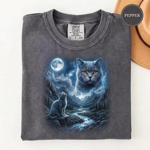 Pode incluir: Uma t-shirt de gola redonda cinzento-carv&atilde;o com uma impress&atilde;o gr&aacute;fica de um gato sentado numa rocha junto a um rio numa paisagem montanhosa sob uma lua cheia, com uma cara de gato grande no c&eacute;u. A etiqueta interior diz Comfort Colors S P CH.