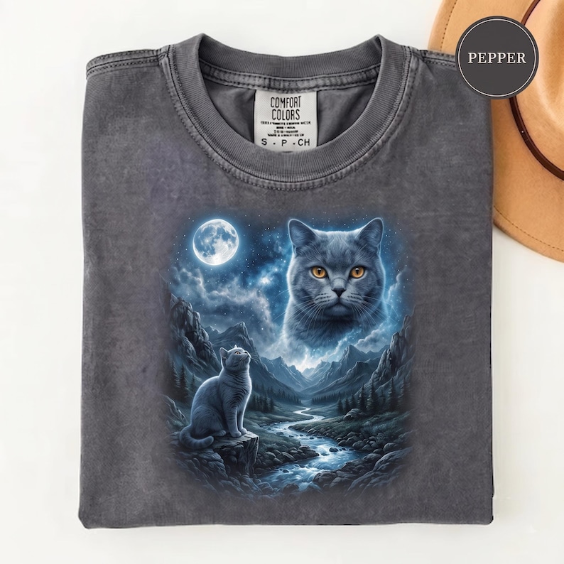 Pode incluir: Uma t-shirt de gola redonda cinzento-carv&atilde;o com uma impress&atilde;o gr&aacute;fica de um gato sentado numa rocha junto a um rio numa paisagem montanhosa sob uma lua cheia, com uma cara de gato grande no c&eacute;u. A etiqueta interior diz Comfort Colors S P CH.