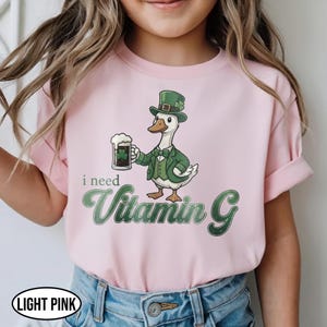 May include: Light pink t-shirt featuring a cartoon duck dressed in a green suit and hat, holding a beer mug with a shamrock. The text "i need Vitamin G" is below the duck. The shirt is labeled "LIGHT PINK".