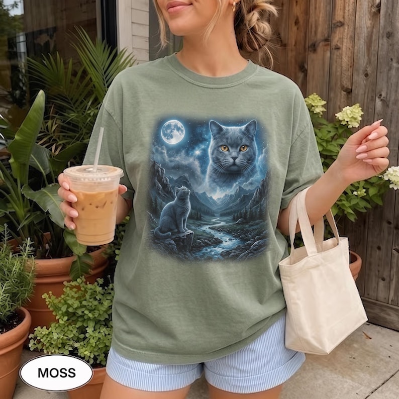 Pode incluir: Uma t-shirt de manga curta verde s&aacute;lvia com uma estampa gr&aacute;fica de um gato sentado numa rocha junto a um rio numa paisagem montanhosa sob a lua cheia, com um rosto de gato fantasmag&oacute;rico no c&eacute;u estrelado acima dos picos das montanhas.