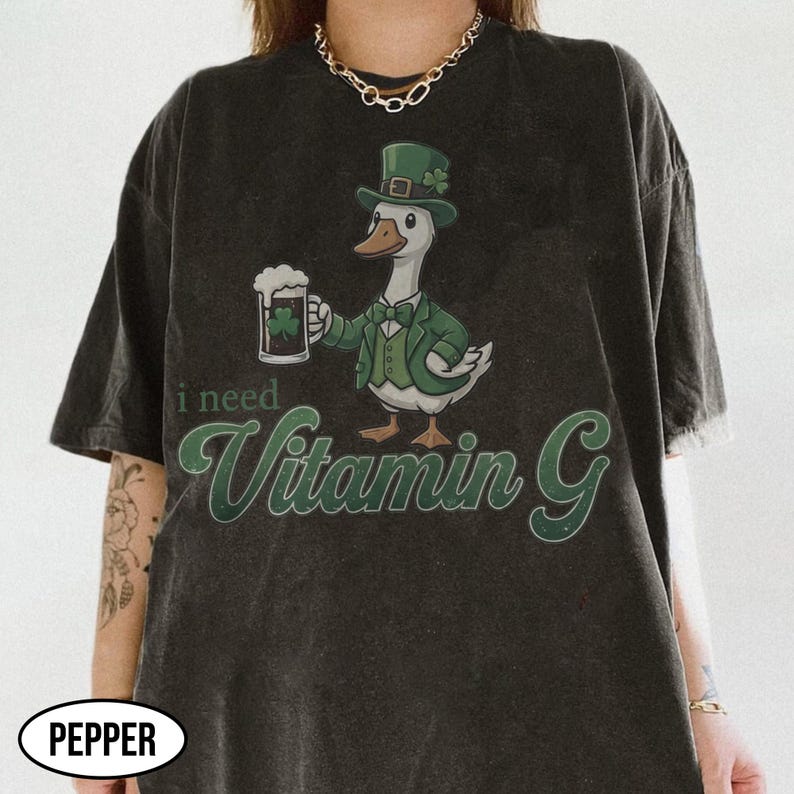 May include: Dark gray t-shirt featuring a cartoon duck dressed in a green suit and hat, holding a beer with a shamrock. The text "i need Vitamin G" is written in green script below the duck.