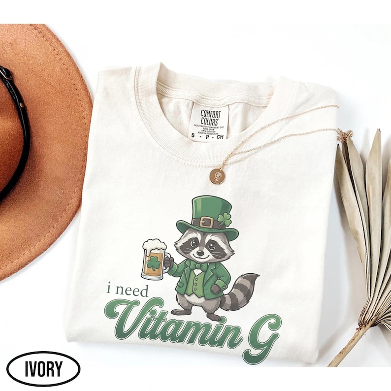 May include: Ivory-colored t-shirt featuring a cartoon raccoon in a green hat and jacket, holding a beer mug with a shamrock. The text "i need Vitamin G" is printed in green. A gold necklace and a brown hat are also visible.