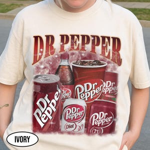 May include: Ivory-colored t-shirt featuring a Dr Pepper graphic. The design includes the Dr Pepper logo, cans, a bottle, and a cup of the soda. The text "Dr Pepper" is prominently displayed in a red, vintage-style font.