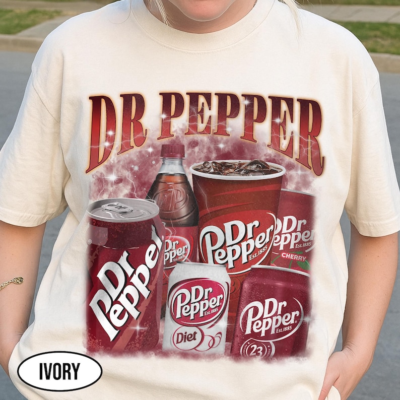 May include: Ivory-colored t-shirt featuring a Dr Pepper graphic. The design includes the Dr Pepper logo, cans, a bottle, and a cup of the soda. The text "Dr Pepper" is prominently displayed in a red, vintage-style font.