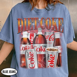 K&ouml;nnte beinhalten: Hellblaues T-Shirt mit einem Diet Coke-Motiv. Das Design zeigt Flaschen, Dosen und einen Becher Diet Coke mit dem Text "DIET COKE" im Retro-Stil. Das T-Shirt hat eine Vintage-&Auml;sthetik.