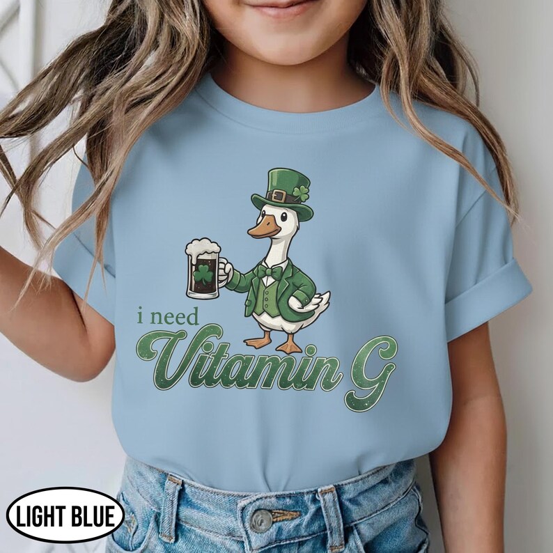 May include: Light blue t-shirt featuring a cartoon goose wearing a green top hat and suit, holding a beer with a shamrock. The text "i need Vitamin G" is in green script.