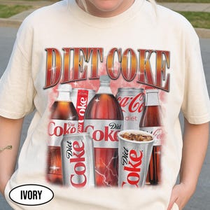 K&ouml;nnte beinhalten: Cremefarbenes T-Shirt mit einem Diet Coke-Motiv. Das Design zeigt verschiedene Diet Coke-Flaschen, Dosen und einen Becher, mit dem Wortlaut "DIET COKE" in fetter, roter und goldener Schrift. Das T-Shirt hat einen Vintage-Look.