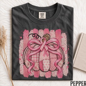 Op de afbeelding: Een antracietgrijs t-shirt met een afbeelding van een pompoen met een roze lint voor borstkankerbewustzijn gestrikt als een strik. De pompoen heeft een patchworkontwerp met diverse roze patronen, waaronder ruitjes, strepen en hartjes, tegen een achtergrond van roze penseelstreken.