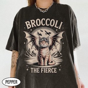 Könnte beinhalten: Dunkelgraues T-Shirt mit einer Grafik einer Katze mit Brille und Fledermausflügeln, mit dem Text "BROCCOLI THE FIERCE". Die Katze ist braun und beige, vor einem Berg- und Himmelshintergrund.