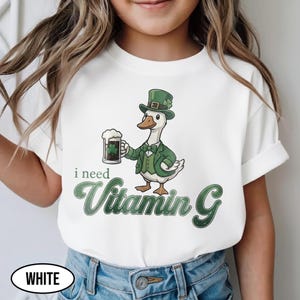 May include: White t-shirt featuring a cartoon goose wearing a green suit and hat, holding a beer. The text "I need Vitamin G" is printed in green. The shirt is a classic crew neck style.