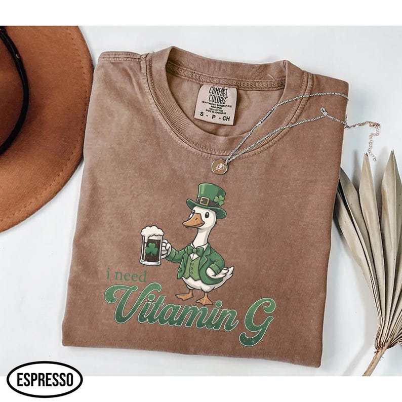 May include: Brown Comfort Colors t-shirt featuring a cartoon goose in a green suit and hat, holding a beer. The text "I need Vitamin G" is printed in green. A silver necklace and a brown hat are also visible.