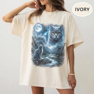 Pode incluir: Uma t-shirt cor de marfim de manga curta com uma estampa gr&aacute;fica de um gato cinzento sentado num penhasco rochoso sobre um riacho de montanha sob a lua cheia, com um grande rosto de gato et&eacute;reo no c&eacute;u noturno estrelado.