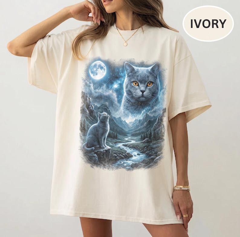 Pode incluir: Uma t-shirt cor de marfim de manga curta com uma estampa gr&aacute;fica de um gato cinzento sentado num penhasco rochoso sobre um riacho de montanha sob a lua cheia, com um grande rosto de gato et&eacute;reo no c&eacute;u noturno estrelado.