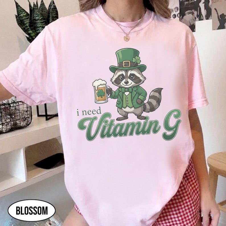 May include: A light pink t-shirt featuring a cartoon raccoon wearing a green hat and jacket, holding a beer mug with a shamrock. The text "i need Vitamin G" is below the raccoon in green script.