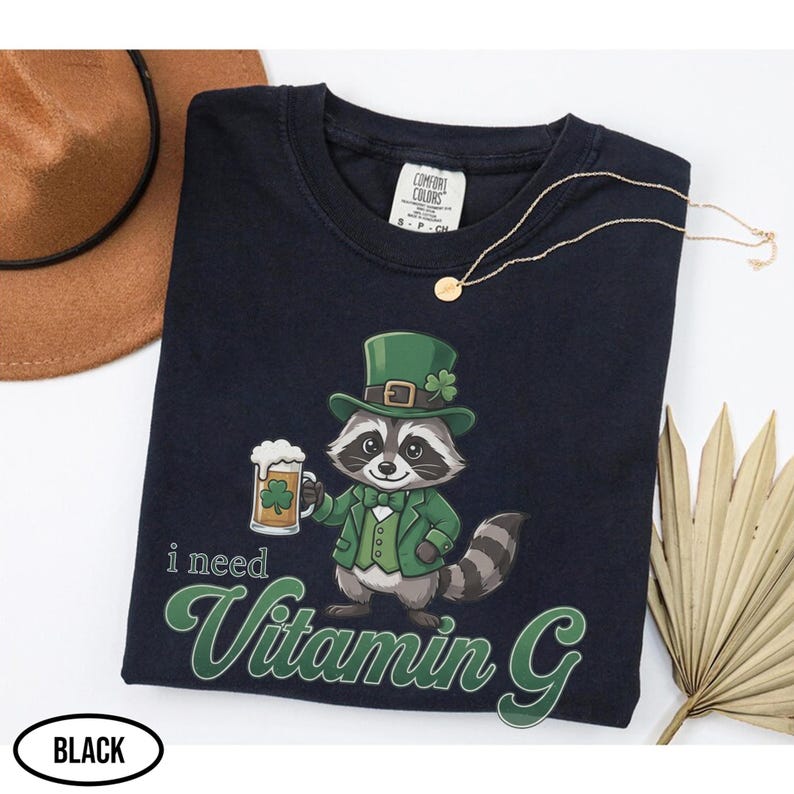 May include: Black t-shirt featuring a cartoon raccoon in a green suit and hat, holding a beer mug with a shamrock. The text "i need Vitamin G" is in green script. A gold necklace and a brown hat are also visible.