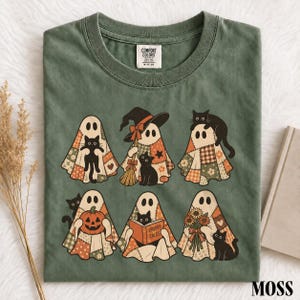 Könnte beinhalten: Ein moosgrünes T-Shirt mit Rundhalsausschnitt und einem Grafikdruck von sechs Geistern in Patchwork-Laken. Die Geister interagieren mit schwarzen Katzen, einem Besen, einem Kürbis, einem Buch mit dem Titel Spooky Tales und Sonnenblumen.