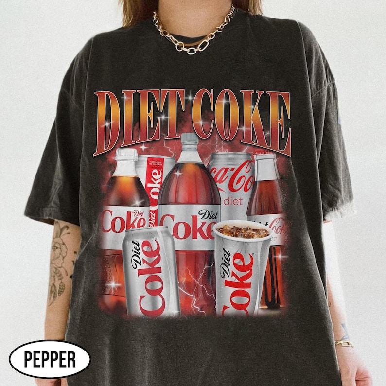 May include: Dark grey t-shirt with a graphic of Diet Coke bottles, cans, and a cup. The text "DIET COKE" is in a red and gold gradient font. The shirt has a vintage, distressed appearance.
