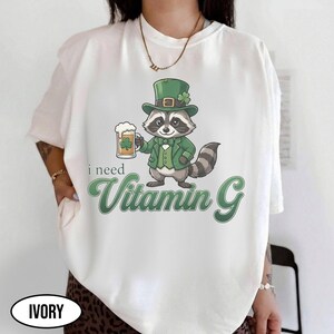 May include: Ivory-colored t-shirt featuring a cartoon raccoon dressed in a green suit and hat, holding a beer mug with a shamrock. The text "i need Vitamin G" is printed in green script below the raccoon.