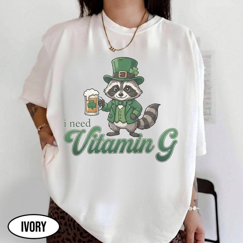 May include: Ivory-colored t-shirt featuring a cartoon raccoon dressed in a green suit and hat, holding a beer mug with a shamrock. The text "i need Vitamin G" is printed in green script below the raccoon.