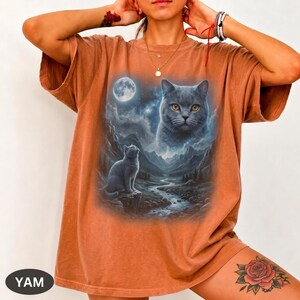 Pode incluir: Uma t-shirt de manga curta cor de ferrugem com uma estampa gr&aacute;fica de um gato sentado num penhasco rochoso sobre um riacho de montanha, com um rosto de gato grande no c&eacute;u noturno acima de uma lua cheia e nuvens estreladas.