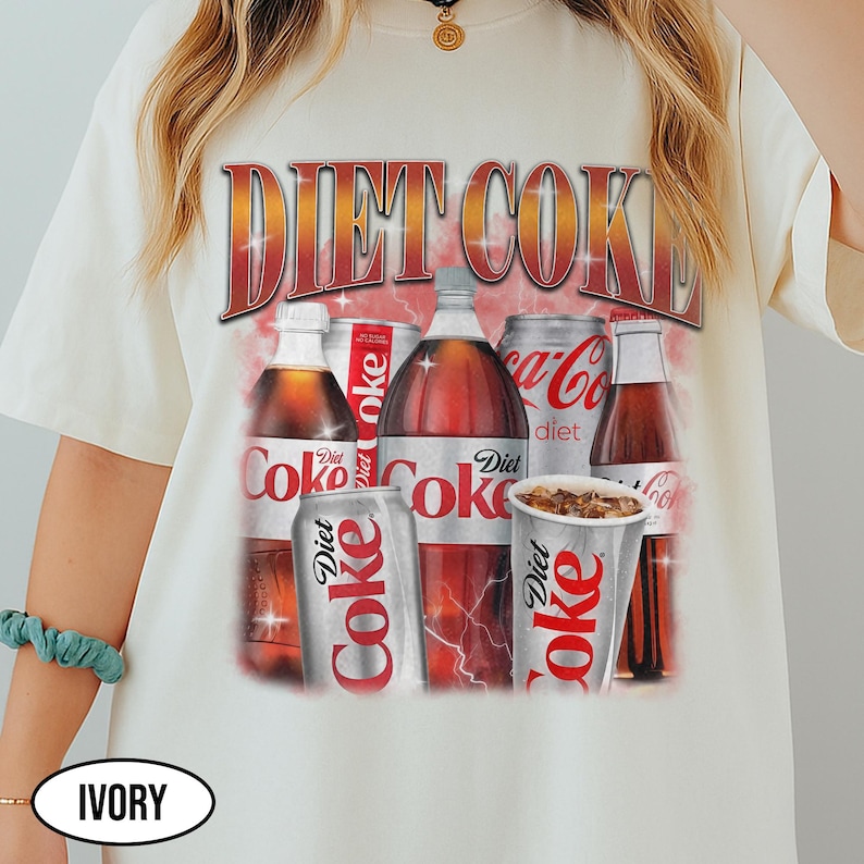 K&ouml;nnte beinhalten: Elfenbeinfarbenes T-Shirt mit einem Diet Coke-Motiv. Das Design zeigt Flaschen, Dosen und einen Becher des Getr&auml;nks, mit dem Schriftzug "DIET COKE" in einem Retro-Stil. Das T-Shirt hat einen Rundhalsausschnitt.