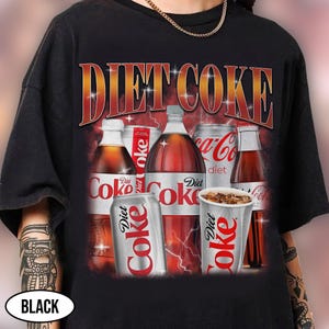 K&ouml;nnte beinhalten: Schwarzes T-Shirt mit einem Diet Coke-Design. Die Grafik zeigt verschiedene Diet Coke-Flaschen, Dosen und einen Becher mit dem Schriftzug "Diet Coke" in einer Retro-Schriftart. Das Design hat ein rotes und oranges Farbschema.
