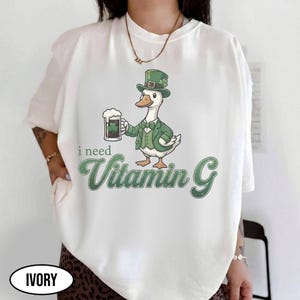 May include: Ivory-colored t-shirt featuring a cartoon duck in a green suit and hat, holding a beer mug with a shamrock. The text "i need Vitamin G" is printed in green, creating a fun, St. Patrick's Day theme.