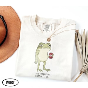 Puede incluir: Sudadera Comfort Colors de color marfil con una rana de dibujos animados sosteniendo una lata de Dr. Pepper. La rana tiene una expresión aburrida, y el texto "I HAVE TO SAY WEIRD STUFF OR I'LL DIE" está impreso debajo. También se ve un collar dorado y un sombrero marrón.