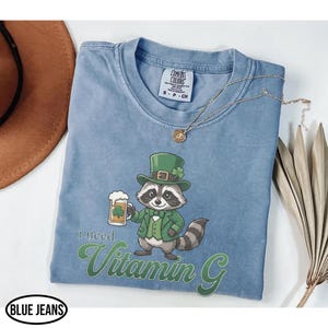 May include: A light blue t-shirt with a graphic of a raccoon wearing a green hat and suit, holding a beer mug with a shamrock. The text "I need Vitamin G" is printed below the raccoon. The shirt has a crew neck and short sleeves.