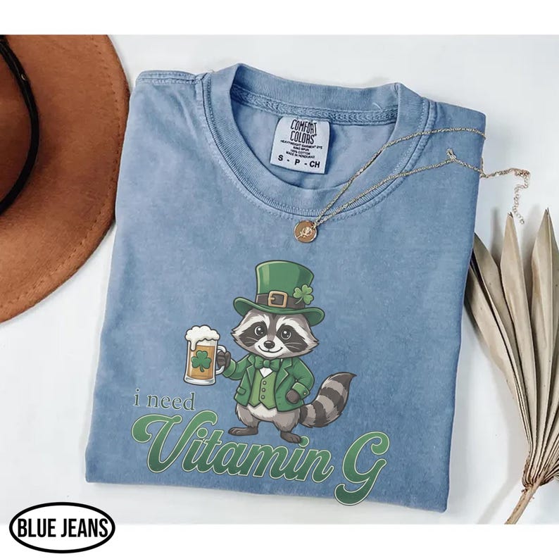 May include: A light blue t-shirt with a graphic of a raccoon wearing a green hat and suit, holding a beer mug with a shamrock. The text "I need Vitamin G" is printed below the raccoon. The shirt has a crew neck and short sleeves.