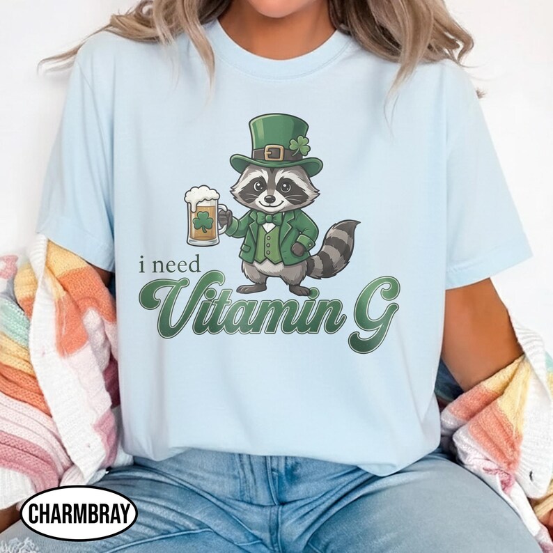 May include: Light blue t-shirt featuring a cartoon raccoon in a green suit and hat, holding a beer mug with a shamrock. The text "i need Vitamin G" is printed in green. The shirt is a casual wear item.