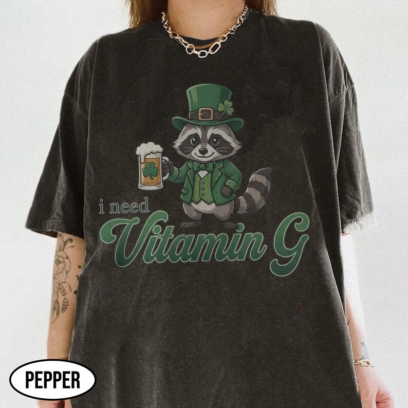 May include: Dark gray t-shirt featuring a cartoon raccoon in a green suit and hat, holding a beer stein with a shamrock. The text "i need Vitamin G" is in green script. The shirt has a distressed look.
