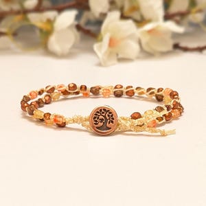 May include: A handmade bracelet with a tree of life charm. The bracelet is made with beige cord and decorated with small, colourful beads in shades of brown, orange, and yellow. The charm is copper-coloured.