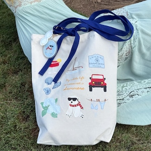 May include: A cream-colored tote bag with a navy blue strap and various embroidered summer-themed designs. The designs include a red jeep, a sailboat, a dog wearing sunglasses, and the text "My whole life measured in summers."